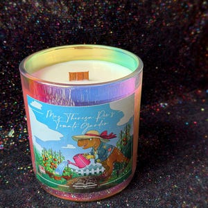 May include: A candle in an iridescent glass jar with a wooden wick. The label features a cartoon dinosaur wearing a sun hat and watering tomato plants with a pink watering can, with the text Mrs. Theresa Rex's Tomato Garden and Story Time Candle Company.