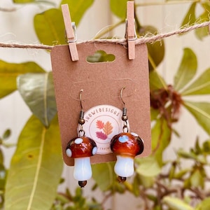 May include: A pair of mushroom-shaped earrings with brown caps, white stems, and bronze-colored hooks. The earrings are displayed on a brown card with the text "HOWEDESIGNS" and a leaf design. The background features green leaves and a twine string.