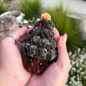 May include: A handmade, plush Christmas tree with a brown base and a green tree top. The tree is decorated with small red accents and topped with a small orange pom-pom. The tree is held in open hands.
