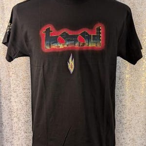 Puede incluir: Una camiseta negra de manga corta con la palabra tool en una fuente estilizada con borde rojo en el pecho. Debajo del texto hay un pequeño gráfico de llama que contiene un ojo central. La camiseta tiene cuello redondo y un diseño de corte estándar.