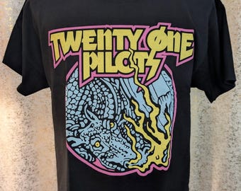 May include: A black short-sleeve t-shirt featuring a graphic print of a blue dragon breathing yellow fire. The text "TWENTY ONE PILOTS" is printed in yellow with a pink outline above the dragon. The design is enclosed within a pink circular border.