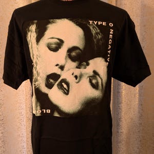 以下が含まれることがあります： 黒の半袖Tシャツで、二つの顔が寄り添うグラフィックプリントが施されています。グラフィックの右側には「TYPE O NEGATIVE」という文字が白で縦にプリントされており、左下には「BLOODY」という単語が一部見えています。