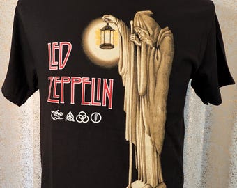 May include: A black short-sleeve t-shirt featuring a graphic of a robed figure holding a lantern while standing on a rocky peak. The text "LED ZEPPELIN" is printed in red with white outlines, accompanied by four circular symbols below the band name.
