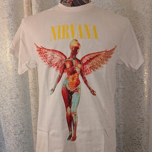 May include: A white short-sleeve t-shirt featuring the yellow text NIRVANA above a graphic of an anatomical human figure with large feathered wings. The figure displays internal organs and skeletal structures in shades of red, orange, and blue.