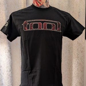 Puede incluir: Una camiseta negra de manga corta con la palabra TOOL impresa en el pecho en una fuente estilizada con contorno blanco. Las letras están rellenas con un patrón complejo de color rojo oscuro y negro. Un pequeño gráfico es visible en la manga derecha.