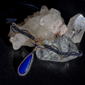 May include: A necklace featuring a teardrop-shaped blue lapis lazuli pendant encased in grey macrame cord. The band consists of dark blue macrame with small gold-toned beads and a central brass lotus flower charm with a small clear stone center.