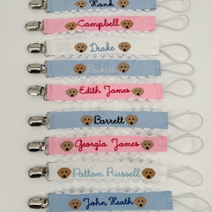 May include: Ten personalized baby bib clips with embroidered names and dog faces. The clips are in blue, pink, and white with scalloped edges. The names are Hank, Campbell, Drake, Beckett, Edith James, Barrett, Georgia James, Patton Russell, and John Heath.