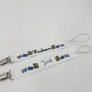 May include: Two white pacifier clips with embroidered train designs in blue and green. One clip says "Hudson" and the other says "Jack" in blue script. Each clip has a silver metal clasp and a white loop.