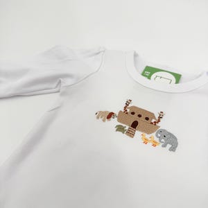 May include: White long-sleeved shirt featuring an embroidered design of Noah's Ark with animals. The design includes a brown ark, a dog, a giraffe, a turtle, a duck, and an elephant. The shirt has a tag attached.