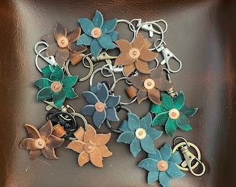 May include: A collection of leather flower keychains in various colors, including brown, green, blue, and gray. The keychains are arranged in a pile on a brown leather surface. Each keychain features a metal ring and a small copper accent in the center of the flower.