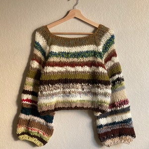 Puede incluir: Un suéter de punto a rayas con cuello amplio de barco y mangas largas. El suéter está hecho de un hilo grueso en tonos de marrón, verde, blanco y crema.