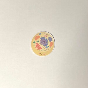 May include: A yellow and white sticker with a cartoon illustration of a cell. The cell has a nucleus, cytoplasm, and other organelles. The cell has a smiling face.