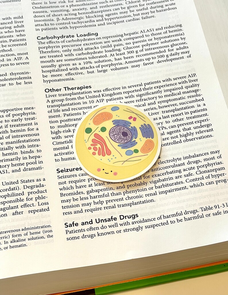 May include: A yellow and orange cell illustration with a smiling face. The cell is on a page of a book with text about porphyria, a rare genetic disorder. The text discusses treatment options, including liver transplantation, and safe and unsafe drugs for patients with porphyria.