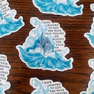 May include: White stickers with a blue wave design and text. The text reads "I have learned to kiss the wave that throws me against the rock of ages." The stickers are on a wooden surface.