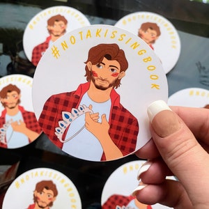 May include: A round sticker with a white background and a yellow border. The sticker features a cartoon illustration of a person with brown hair and a red and black plaid shirt. The person is holding a crown in their right hand. The text "#NOTAKISSINGBOOK" is written in yellow letters around the border of the sticker.