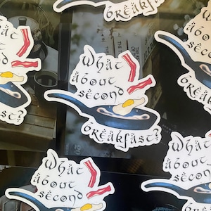Op de afbeelding: Vier stickers met een blauwe koekenpan, een gebakken ei, spek en de tekst "What about second breakfast?" in een gestileerd lettertype.