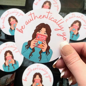 May include: Round sticker with a white background and pink text that says "be authentically you". The sticker features a cartoon illustration of a woman with long brown hair holding a book that says "Authentically Izzy".