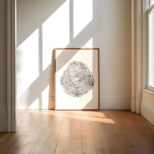 Könnte beinhalten: Ein gerahmter Druck eines Baumquerschnitts. Das Kunstwerk zeigt eine detaillierte Schwarz-Weiß-Illustration von Baumringen, die in einem hellbraunen Holzrahmen eingefasst ist. Der Druck lehnt an einer weißen Wand, mit Sonnenlicht.