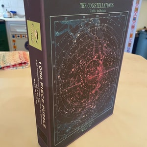 May include: A boxed 1000-piece jigsaw puzzle featuring a detailed star chart of constellations visible in Britain. The box is dark purple with gold lettering and a colorful illustration of the night sky. The puzzle measures 48 x 69 cm and is made in the UK.