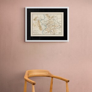 May include: A framed vintage map of the city of Ancient Rome, with a black frame and a white mount, hanging on a pale pink wall. A wooden chair is in the foreground.