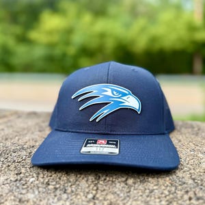 May include: Navy blue baseball cap with a light blue and white eagle logo on the front. The cap has a curved brim and a structured fit. A small tag is visible on the front. The background is blurred green.