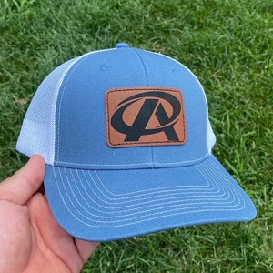 Puede incluir: Una gorra de camionero azul y blanca con un parche de cuero marrón que presenta una letra "A" estilizada en negro.