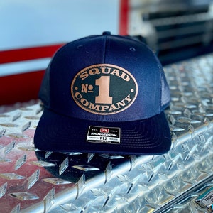 May include: Navy blue trucker hat with a mesh back and a curved brim. The front features a brown oval patch with the words "SQUAD COMPANY" and the number "1". The hat is adjustable and has a Richardson tag.