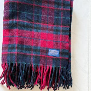 May include: A folded red, black, and blue plaid blanket with a fringed edge. The blanket has a rectangular blue tag with white text. The fringe is a mix of red and blue. The blanket is made of a soft material.
