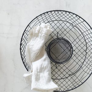 May include: A black wire basket with a circular design, featuring a smaller wire mesh basket inside. A white linen cloth is draped over the edge of the basket. The items are set against a white marble background.