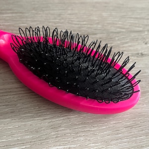 May include: A bright pink oval hairbrush with black bristles. The brush has a curved handle for easy grip and is designed for detangling and styling hair. The bristles are made of a flexible material.