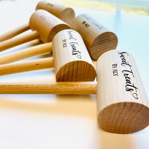 May include: A collection of wooden mallets with rounded heads and long handles. The mallets are light brown and feature the text "Sweet Treats BY ALY" in black script. The mallets are arranged on a white surface.