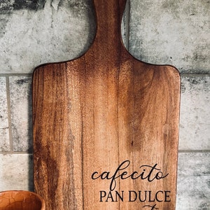 May include: A wooden chopping board with a handle. The board features the words "cafecito, pan dulce y tu" in script. The wood grain is visible, and the board is a warm brown colour. The board is rectangular.