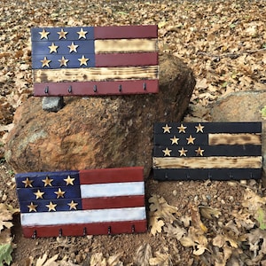 May include: Three wooden American flag-themed key holders. Each features a distressed finish with painted red, white, and blue stripes and gold star accents. The key holders have small hooks along the bottom edge.