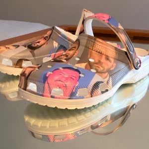 Op de afbeelding: Witte klompen met een kleurrijke collage van gezichten die op de voorkant zijn gedrukt. De gezichten zijn van verschillende mensen, sommige met een serieuze uitdrukking en andere met een meer speelse uitdrukking.