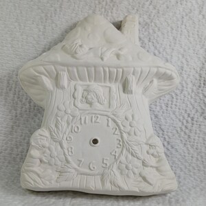 May include: White ceramic clock shaped like a mushroom house with a clock face and a small gnome figure on the side. The clock face has Roman numerals and a hole in the center.