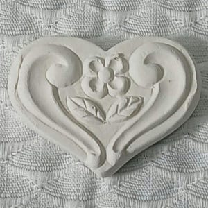 May include: White ceramic heart-shaped ornament with a raised floral design. The flower has five petals and is surrounded by two leaves.