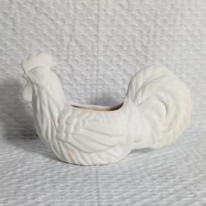 Może przedstawiać: Biała ceramiczna doniczka w kształcie koguta z detalicznym wzorem. Doniczka ma pustą przestrzeń w środku do sadzenia.