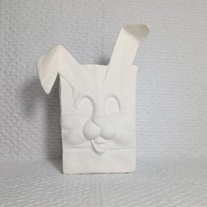 Könnte beinhalten: Ein weißer Keramikbehälter in Form eines Osterhasen. Der Behälter hat eine rechteckige Form mit einem Hasengesicht-Design auf der Vorderseite, einschließlich Ohren, Augen, Nase und einem lächelnden Mund. Die Ohren ragen über die Oberseite des Behälters hinaus.