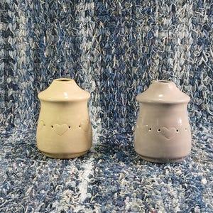 May include: Two ceramic candle warmers with heart cutouts. The warmer on the left is beige and the warmer on the right is light gray.