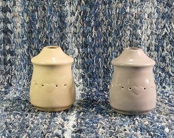 May include: Two ceramic candle warmers with heart cutouts. The warmer on the left is beige and the warmer on the right is light gray.