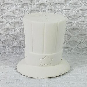 May include: A white ceramic top hat with a star on the band. The hat has vertical lines and a wide brim. The hat is a decorative item.