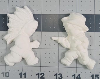 May include: Two white ceramic figurines, one depicting a person wearing a headdress with feathers and the other a person wearing a hat. Both figurines are standing and facing forward.
