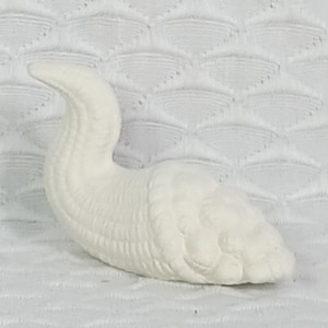 May include: A white ceramic swan figurine with a curved neck and detailed feather texture. The swan is resting on a textured white surface. The figurine is a decorative item.