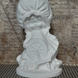 May include: White ceramic figurine of a girl with a bonnet spinning a wheel. The figurine is in a sitting position and has a basket of yarn at her feet.