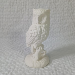 Pode incluir: Figurinha de coruja de cerâmica branca com penas detalhadas e uma pose empoleirada. A coruja está empoleirada em um galho com uma base.