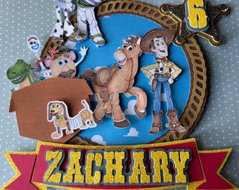 May include: A colorful cake topper featuring characters from the Toy Story movie. The topper includes Woody, Buzz Lightyear, Jessie, and other characters, with the name "ZACHARY" and the number "6" displayed.