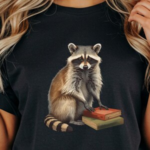 May include: Black t-shirt featuring a watercolor illustration of a raccoon sitting on two books. The raccoon has gray and brown fur with a striped tail. The books are red and green.