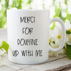 May include: White ceramic mug with the text "MERCI FOR POUTINE UP WITH ME" in black script. The mug is set against a blurred background of greenery and white flowers.