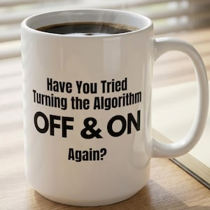 May include: A white ceramic mug with a handle, featuring black printed text that reads: Have You Tried Turning the Algorithm OFF & ON Again? The mug is filled with a dark brown liquid, likely coffee, and has a smooth, cylindrical body shape.