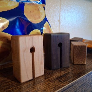 Puede incluir: Tres bloques rectangulares de madera de diferentes tamaños y tonos, cada uno con una ranura vertical y un corte circular en la parte superior, diseñados para sostener objetos en posición vertical. Los bloques están dispuestos en fila, mostrando acabados de madera clara, oscura y natural.